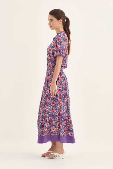 A woman modeling a short-sleeved blouse and a skirt by Verge with a purple, red, and blue floral pattern, shown from a side profile against a plain off-white studio background.