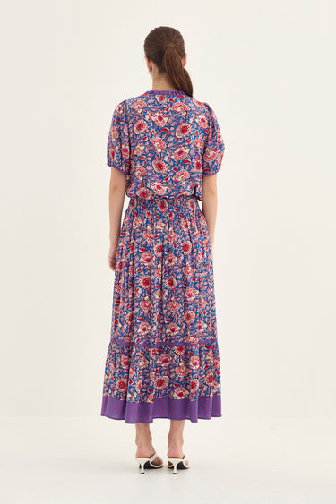 A woman viewed from the back wearing a short-sleeved blouse and a skirt by Verge with a purple, red, and blue floral pattern, set against a plain off-white studio background.