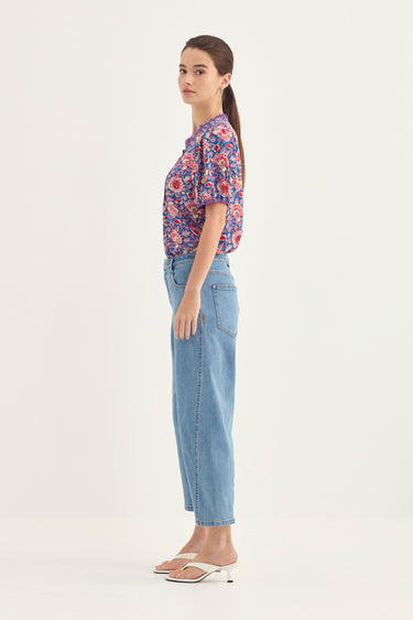 A woman stands in profile wearing a short-sleeve button-up blouse with a dense floral print in purple, pink, and blue, paired with light-wash cropped blue jeans by Verge and white heeled sandals, against a plain off-white studio background.