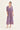 A woman modeling a purple floral dress by Verge with three-quarter sleeves and a V-neckline, paired with white heeled sandals, against a plain off-white studio background.