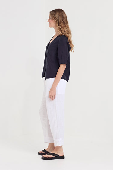 A woman shown in profile wearing a dark navy short-sleeve top, white cropped pants by Verge, and black flip-flop sandals against a plain light gray studio background.