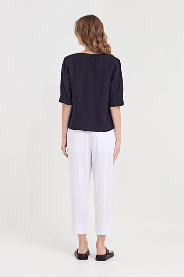 A woman shown from the back wearing a dark navy short-sleeve blouse and white cropped trousers by Verge, standing against a plain light gray studio background.