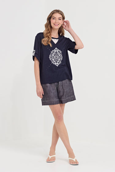 A woman modeling a navy blue short-sleeve t-shirt with a white geometric pattern on the front, paired with gray plaid shorts by Verge and white flip-flop sandals, against a plain white studio background.