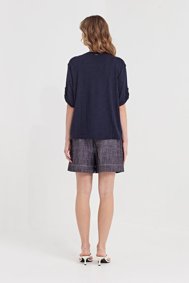 A woman viewed from the back wearing a loose navy short-sleeve top and textured gray shorts by Verge, standing against a plain light gray studio background.
