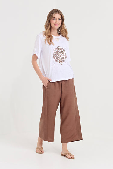 A woman modeling a white short-sleeve t-shirt with a brown mandala print and wide-leg brown pants by Verge, standing against a light gray studio background.