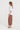 A woman stands in profile wearing a loose white short-sleeve T-shirt and brown wide-leg cropped pants by Verge, paired with tan sandals, against a plain light gray studio background.