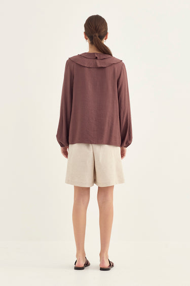 A woman viewed from the back wearing a loose, long-sleeve mauve-brown top with a wide collar, light beige shorts, and black sandals by Verge, standing against a plain off-white studio background.