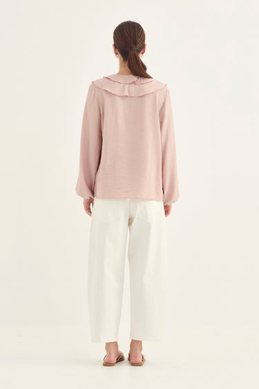 A woman viewed from the back wearing a dusty pink long-sleeve top with a wide ruffled collar and white wide-leg cropped pants by Verge, standing against a plain light background.