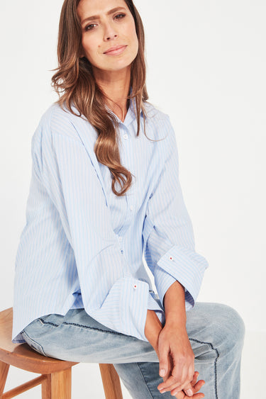 Woman wearing a light blue striped shirt and jeans by Verge sitting on a wooden stool against a white background