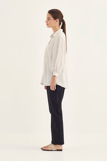 A woman shown in profile wearing a cream and dark vertical-striped button-up shirt, dark navy pants by Verge, and beige slide sandals, standing against a plain off-white studio background.