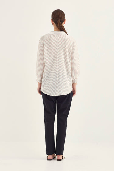 A woman viewed from the back wearing a cream-colored long-sleeve blouse with a fine diagonal striped pattern and dark navy pants by Verge, standing against a plain off-white studio background.