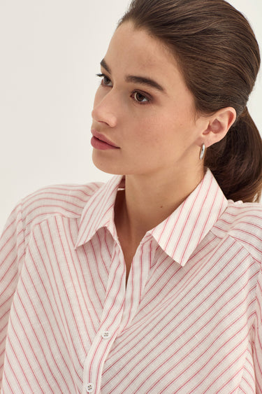 A close-up of a woman wearing a pink and white vertically striped button-down shirt by Verge, shown from the chest up against a plain light background.