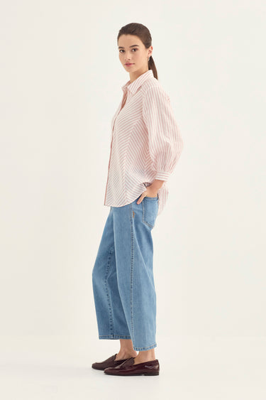 A woman modeling a light pink and white vertically striped button-down shirt with voluminous sleeves, paired with wide-leg light blue jeans by Verge and dark brown loafers, set against a plain off-white studio background.