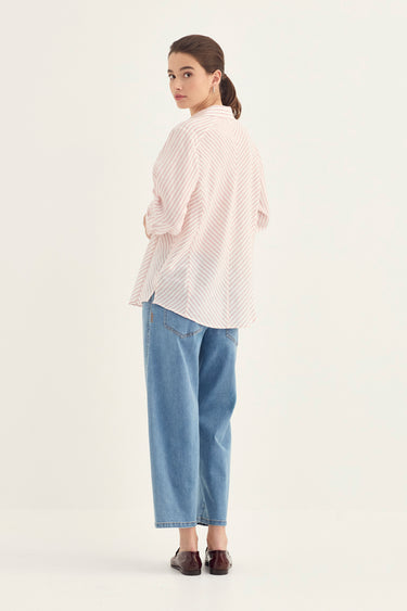 A woman stands in profile wearing a light pink and white striped button-up shirt, light blue cropped jeans by Verge, and dark brown loafers against a plain off-white studio background.