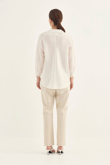 A woman viewed from the back wearing a cream-colored textured long-sleeve blouse and beige trousers by Verge, standing against a plain off-white studio background.