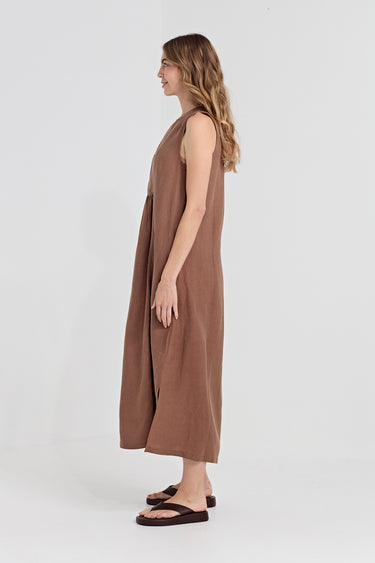 A woman stands in profile wearing a sleeveless, loose-fitting midi dress by Verge in a warm taupe-brown color, paired with dark brown flip-flop sandals, against a plain light gray studio background.
