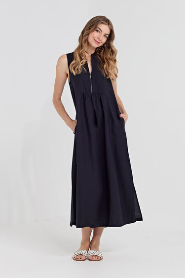 A woman modeling a sleeveless navy midi dress by Verge with a front zipper, side pockets, and a gathered waist, paired with silver embellished flat sandals, against a light gray studio background.