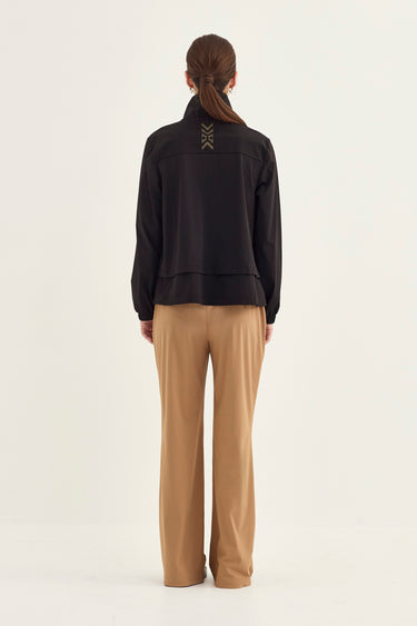 A model shown from the back wearing a black cropped zip-up jacket with lace-up detailing at the back of the neck and beige wide-leg trousers by Verge, set against a plain light gray studio background.
