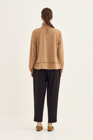 A woman viewed from the back wearing a tan long-sleeve cropped jacket with lace-up detailing on the upper back, paired with black pants and tan flats by Verge, against a plain light background.