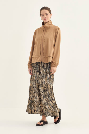 A model stands against a plain light background wearing a tan long-sleeve mock-neck top and a long pleated skirt by Verge with a beige, brown, and black snake print.