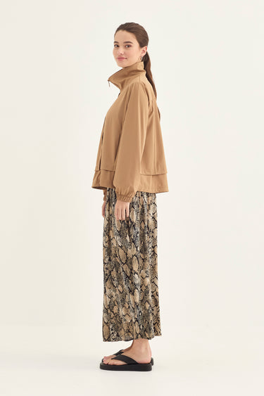 A woman stands in profile wearing a tan cropped jacket with a high collar, a long patterned skirt by Verge, in beige, brown, and gray tones, and black flip-flop sandals against a plain light background.