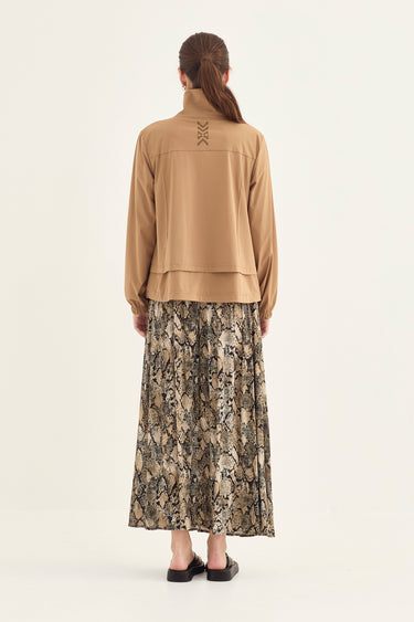 A model shown from the back wearing a tan long-sleeve cropped jacket with lace-up detailing on the back collar and a long pleated skirt by Verge. with a beige, brown, and black snakeskin print, set against a plain off-white background.