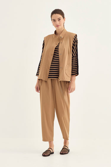 A model stands against a plain light background wearing a tan sleeveless vest over a black and tan striped long-sleeve top, paired with matching tan wide-leg pants and black woven sandals by Verge.