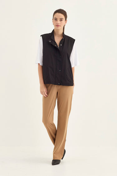 A woman modeling a sleeveless black button-front vest over a white short-sleeve shirt and tan straight-leg pants by Verge, photographed against a plain light background.