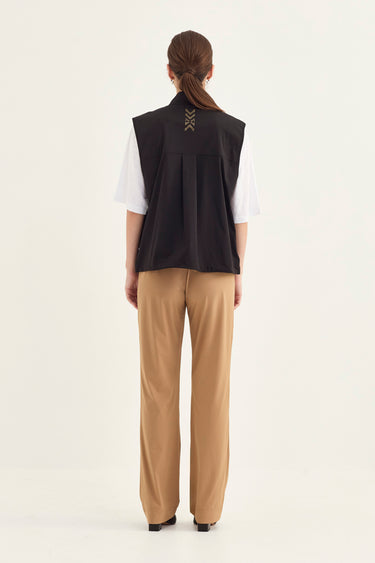 A model shown from the back wearing a black sleeveless vest over a white short-sleeve shirt and beige wide-leg trousers by Verge, against a plain off-white studio background.