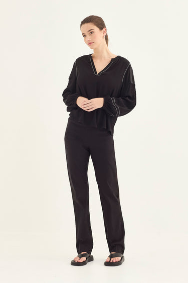 A woman modeling a black lounge set consisting of a long-sleeve V-neck top and matching straight-leg pants by Verge, paired with black sandals, against a plain off-white studio background.