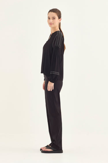A woman stands in profile wearing a black long-sleeve top with thin vertical striped details on the upper sleeves and black wide-leg pants by Verge, photographed against a plain light background.