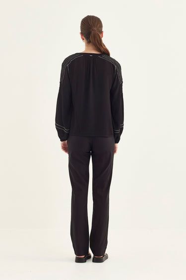 A woman viewed from the back wearing a black long-sleeve top with thin white contrast piping and black trousers by Verge, standing against a plain white studio background.