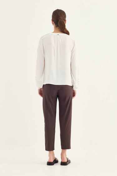 A woman viewed from behind wearing a white long-sleeve blouse and dark brown cropped trousers by Verge, standing against a plain light background.