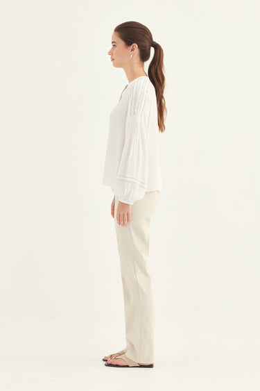 A woman shown in side profile wearing a white blouse and beige pants by Verge against a plain light gray studio background.
