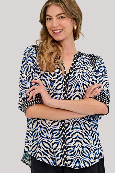 A woman wearing a blue, black, and white abstract patterned blouse by Verge with three-quarter sleeves, standing with arms crossed against a solid gray background.