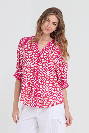 A woman modeling a vibrant pink and white patterned blouse with three-quarter sleeves, paired with white pants by Verge, against a plain light gray background.