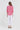 A woman viewed from the back wearing a bright pink and white patterned blouse with three-quarter sleeves and white cropped pants by Verge, standing against a plain light gray studio background.