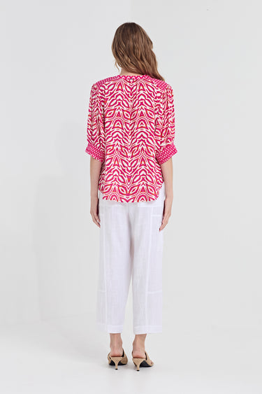 A woman viewed from the back wearing a bright pink and white patterned blouse with three-quarter sleeves and white cropped pants by Verge, standing against a plain light gray studio background.