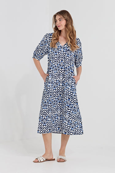 A woman modeling a short-sleeve midi dress by Verge with a blue, white, and black wavy abstract pattern, standing against a solid turquoise background.
