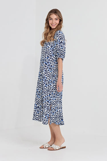 A woman modeling a blue and white print midi dress by Verge  with short sleeves and a side slit, paired with white slide sandals, against a light gray studio background.