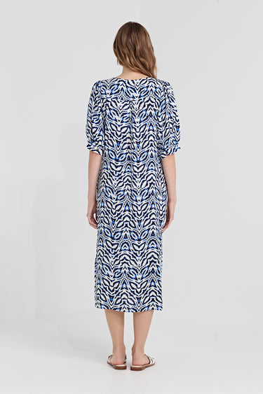 A woman viewed from the back wearing a knee-length dress by Verge  with a blue and white wavy abstract pattern, short puff sleeves, and a round neckline, standing against a light gray studio background.