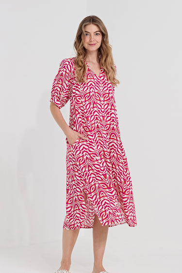 A woman modeling a pink and white wavy patterned midi dress by Verge  with short puff sleeves, a V-neckline, and side pockets, set against a plain light gray studio background.