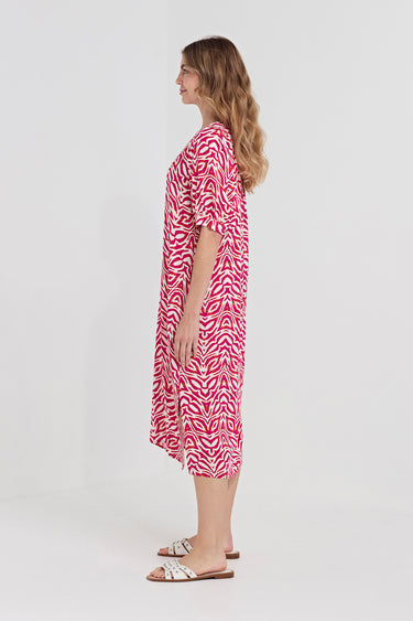 A woman stands in profile wearing a loose, short-sleeve midi dress by Verge with an allover pink and white abstract wavy pattern, paired with white slide sandals, against a plain light gray studio background.