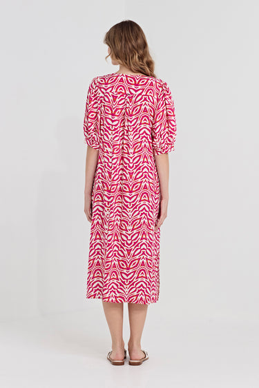 A woman viewed from the back wearing a pink and white patterned midi dress by Verge with short puff sleeves and a gathered waist, paired with flat sandals, against a plain white studio background.