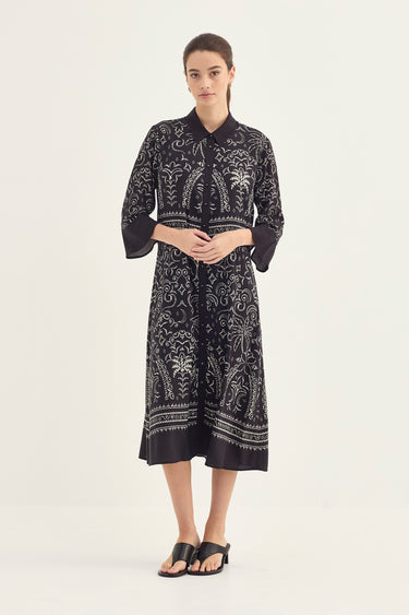 A woman modeling a black midi shirt dress by Verge with a white botanical and paisley print, featuring three-quarter bell sleeves and a button-up fron, set against a plain light gray studio background.