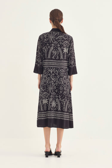 A woman viewed from the back wearing a black dress by Verge with an intricate white floral print, set against a plain light gray studio background.