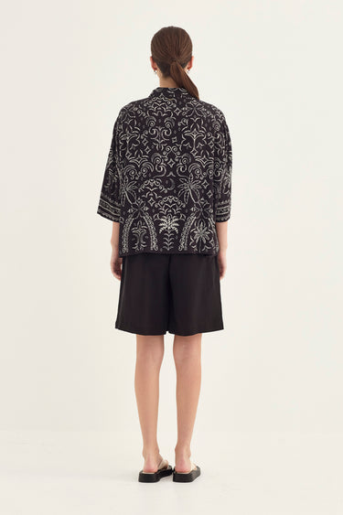 A woman viewed from the back wearing a black short-sleeve top with an intricate white floral or paisley-style print, paired with black shorts by Verge and black flat sandals, against a plain light background.