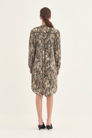 A woman viewed from behind wearing a long-sleeve midi dress by Verge with a beige, brown, and black snakeskin pattern, paired with black pointed-toe flats, against a plain off-white studio background.
