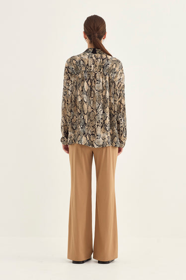 A woman viewed from the back wearing a long-sleeve snakeskin print blouse and tan wide-leg trousers by Verge, standing against a plain light background.