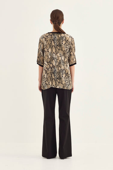 A woman viewed from the back wearing a short-sleeve top with a beige and black snakeskin pattern and black wide-leg pants by Verge, against a plain light background.